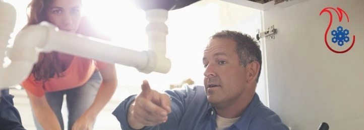 Technician inspecting plumbing pipes under a sink while explaining repairs to a homeowner.